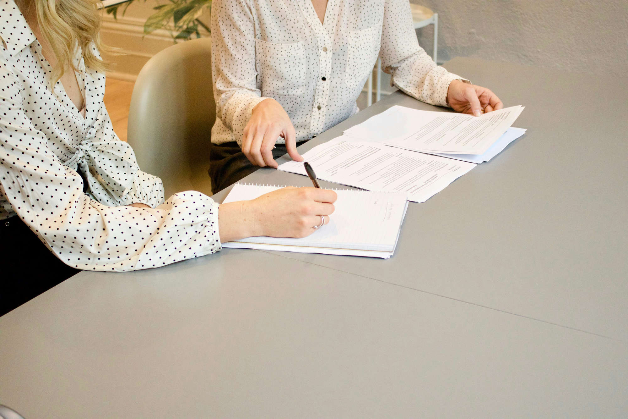 Two individuals in patterned blouses sitting at a table, writing notes and reviewing printed documents.