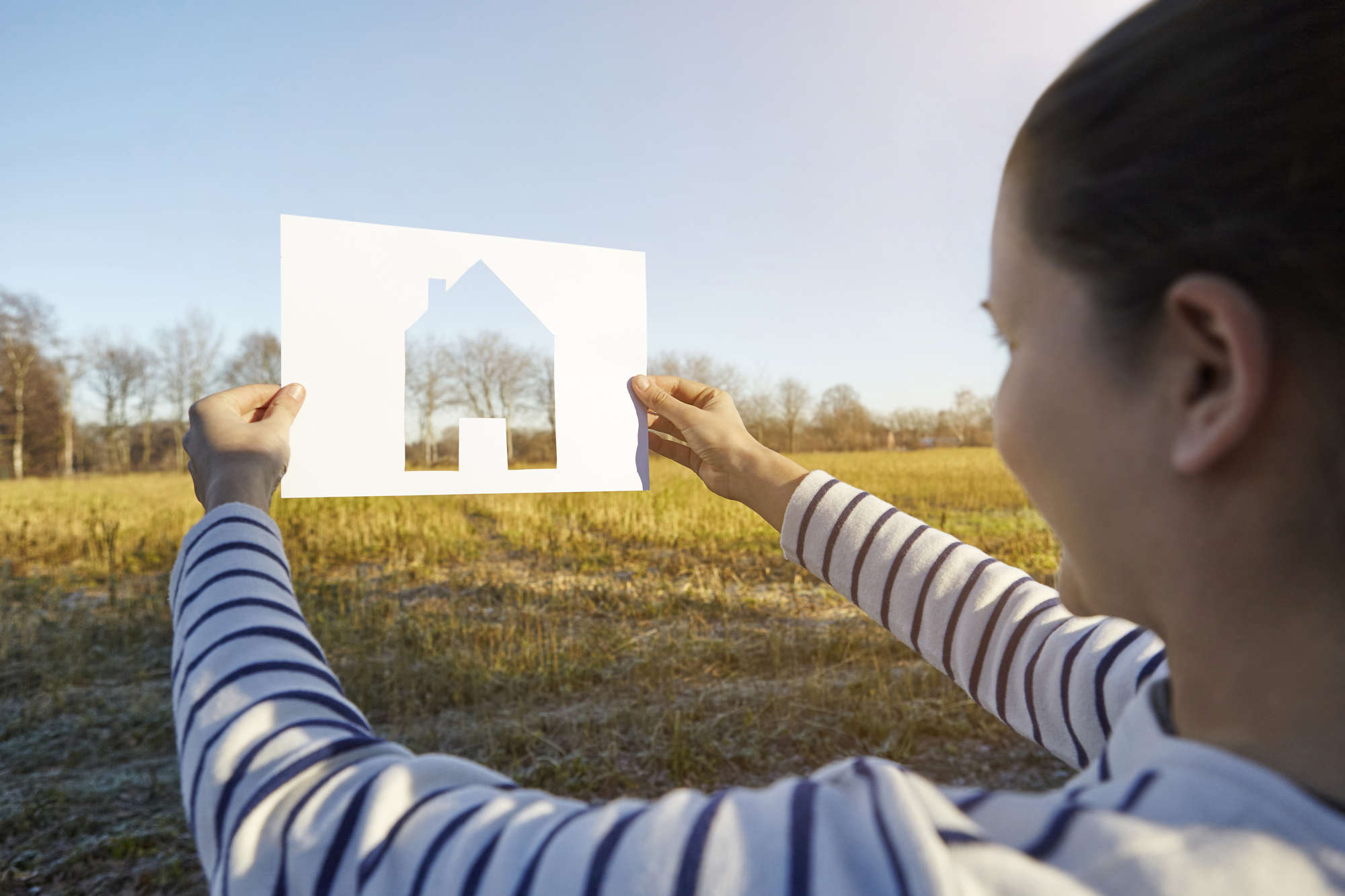 Woman Standing In Rural Setting, Holding Out Paper With Cut Out Of House